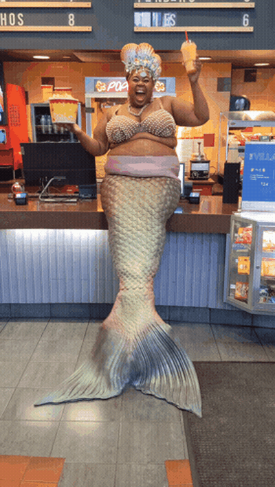 A Woman Dressed As A Mermaid Holds A Drink In Front Of A Pop Corn Machine GIF