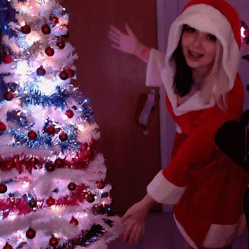 A Woman Dressed In A Santa Claus Outfit Stands In Front Of A Christmas Tree GIF