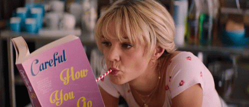 A Woman Drinking From A Straw While Reading A Book Titled Careful How You Go GIF