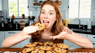 A Woman Eating A Chocolate Chip Cookie From A Plate Of Cookies GIF