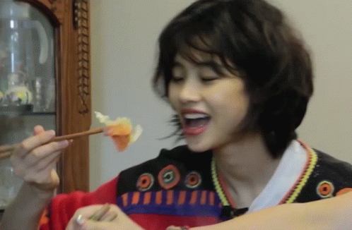 A Woman Eating A Piece Of Food With Chopsticks While Wearing A Sweater With Circles On The Front GIF