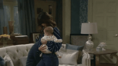 A Woman Holding A Baby In Her Arms In A Living Room GIF