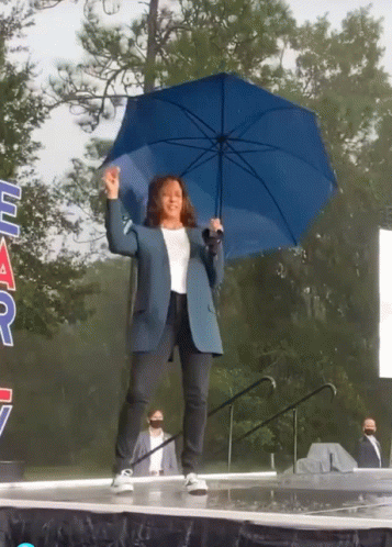 A Woman Holding A Blue Umbrella Stands On A Stage In Front Of A Sign That Says " Har " GIF