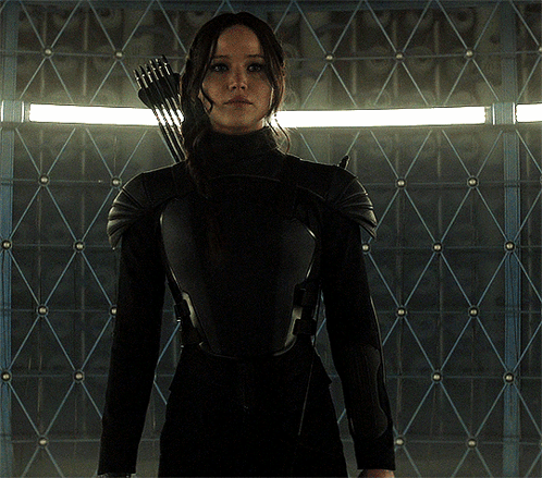 A Woman Holding A Bow And Arrow Stands In Front Of A Wall GIF