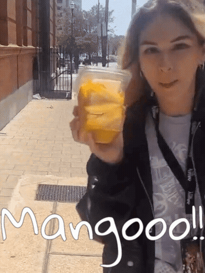 A Woman Holding A Cup Of Mango Juice With The Words Mangooo Written On The Sidewalk GIF