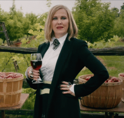 A Woman Holding A Glass Of Wine In Front Of Baskets Of Fruit GIF