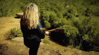 A Woman Holding A Guitar Standing On A Cliff Overlooking A River GIF