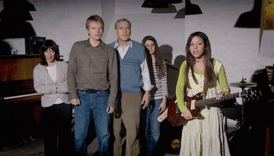 A Woman Holding A Guitar Stands In Front Of A Group Of People GIF