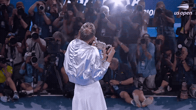 A Woman Holding A Trophy In Front Of A Crowd With Us Open On The Screen GIF