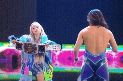 A Woman Holding A Wrestling Belt Stands Next To Another Woman Without A Shirt On GIF