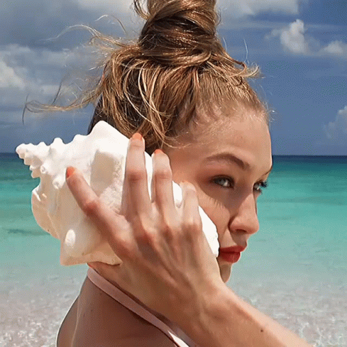 A Woman Holds A Sea Shell To Her Ear On A Beach GIF