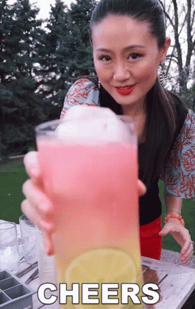 A Woman Holds Up A Glass Of Pink And Yellow Liquid With The Words Cheers Written Below Her GIF