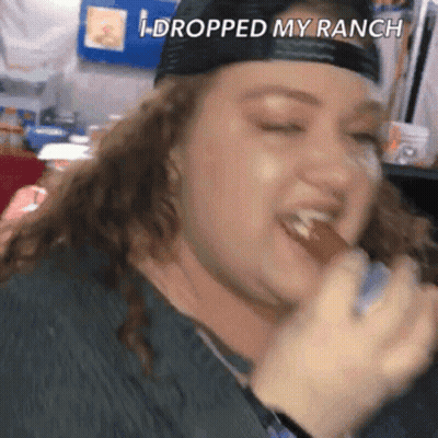 A Woman In A Baseball Cap Is Eating A Hot Dog With The Words I Dropped My Ranch Above Her . GIF
