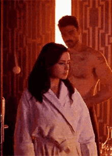 A Woman In A Bathrobe Is Standing Next To A Shirtless Man In A Bathroom . GIF