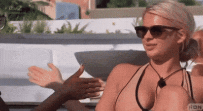 A Woman In A Bikini And Sunglasses Is Sitting On A Couch Talking To Another Woman . GIF