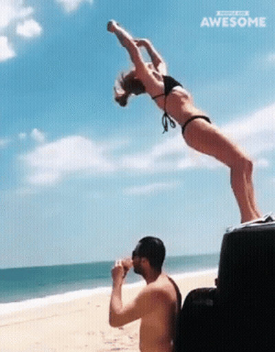 A Woman In A Bikini Is Doing A Handstand While A Man Drinks From A Cooler On The Beach GIF