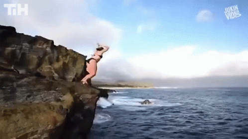 A Woman In A Bikini Is Jumping Off Of A Cliff Into The Ocean . GIF