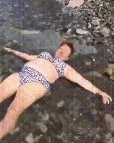 A Woman In A Bikini Is Laying On A Rocky Beach With Her Arms Outstretched . GIF