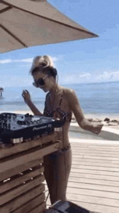 A Woman In A Bikini Is Standing In Front Of A Dj Machine On The Beach . GIF