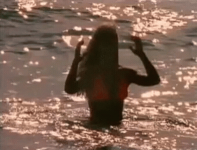 A Woman In A Bikini Is Standing In The Ocean At Sunset . GIF