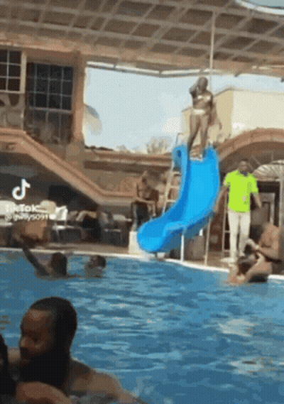 A Woman In A Bikini Is Standing On Top Of A Blue Water Slide In A Swimming Pool . GIF