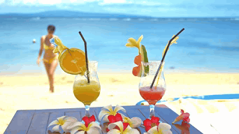 A Woman In A Bikini Is Walking On The Beach Behind Two Drinks With Straws GIF