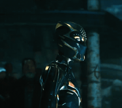A Woman In A Black And Gold Costume With A Helmet On Stands In A Dark Room GIF