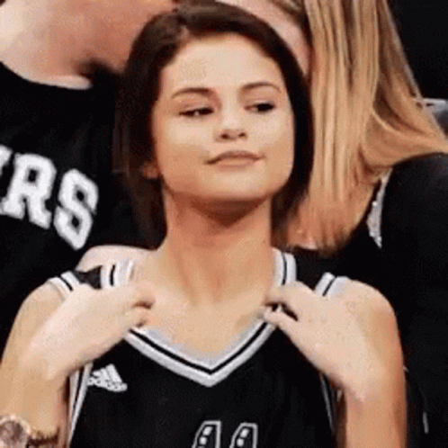 A Woman In A Black And White Basketball Jersey Is Sitting In A Crowd Of People . GIF