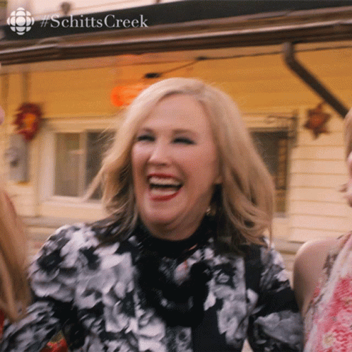 A Woman In A Black And White Floral Dress Is Laughing In Front Of A Sign That Says Schitts Creek GIF
