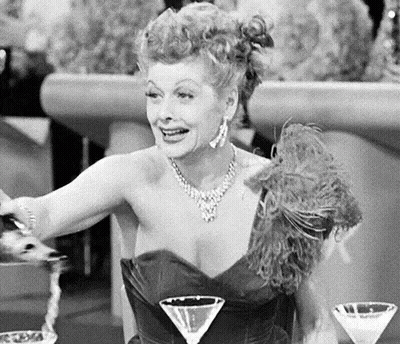 A Woman In A Black And White Photo Is Sitting At A Table With Two Martini Glasses . GIF