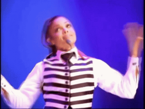 A Woman In A Black And White Striped Jacket Is Singing Into A Microphone . GIF