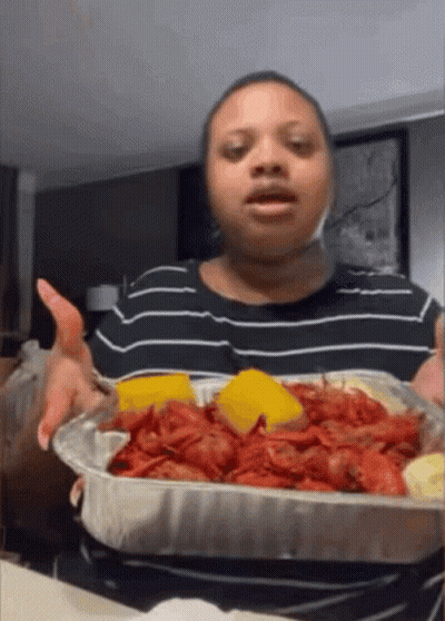 A Woman In A Black And White Striped Shirt Is Holding A Tray Of Crawfish And Cornbread GIF