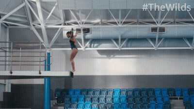 A Woman In A Black Bathing Suit Is Jumping Off A Diving Board With #Thewilds On The Bottom GIF