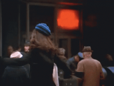 A Woman In A Black Coat And Hat Is Standing In Front Of A Red Light . GIF