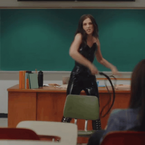 A Woman In A Black Corset Is Standing In Front Of A Chalkboard In A Classroom GIF