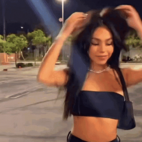 A Woman In A Black Crop Top Is Adjusting Her Hair In A Parking Lot . GIF