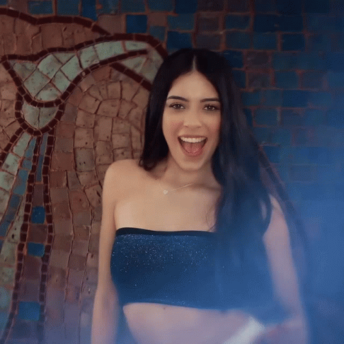 A Woman In A Black Crop Top Is Smiling In Front Of A Mosaic Wall GIF