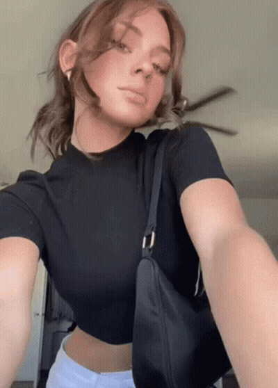 A Woman In A Black Crop Top Is Taking A Selfie While Holding A Black Bag . GIF