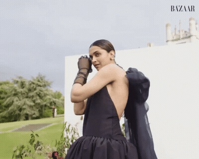 A Woman In A Black Dress And Black Gloves Is Standing In Front Of A White Wall That Says Bazaar On It GIF
