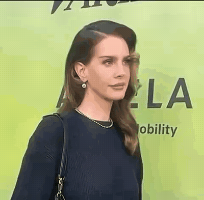 A Woman In A Black Dress And Pearl Earrings Is Standing In Front Of A Green Background . GIF