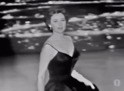 A Woman In A Black Dress And White Gloves Is Dancing On A Stage . GIF