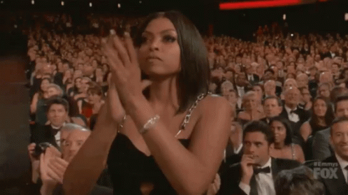 A Woman In A Black Dress Is Applauding In Front Of A Crowd Of People . GIF