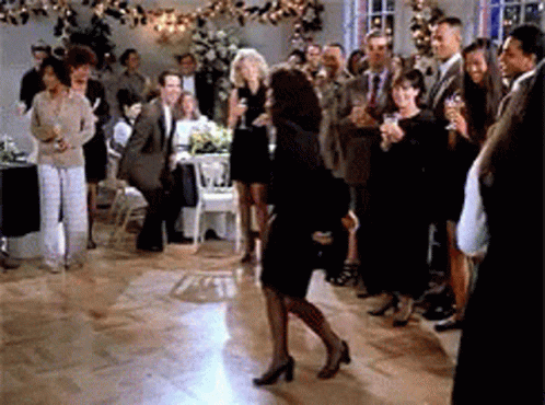 A Woman In A Black Dress Is Dancing In Front Of A Crowd At A Party GIF