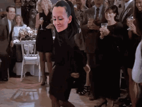 A Woman In A Black Dress Is Dancing In Front Of A Crowd Of People At A Party . GIF