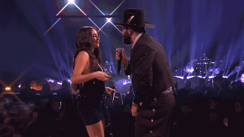 A Woman In A Black Dress Is Dancing On A Stage With A Man In A Cowboy Hat GIF