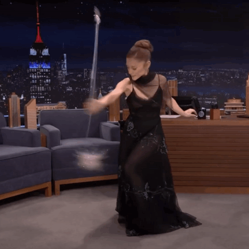 A Woman In A Black Dress Is Dancing With A Wand In Front Of The Empire State Building GIF