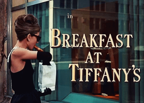 A Woman In A Black Dress Is Holding A Bag Of Food In Front Of A Sign That Says Breakfast At Tiffany 'S GIF