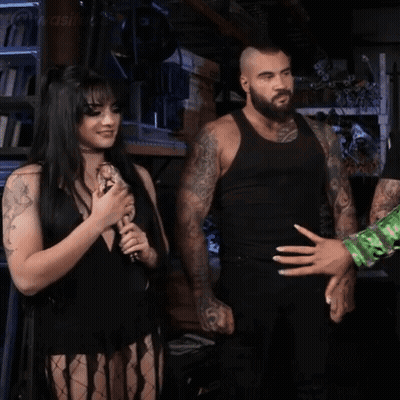 A Woman In A Black Dress Is Holding A Microphone And Smiling While A Man Stands Behind Her . GIF