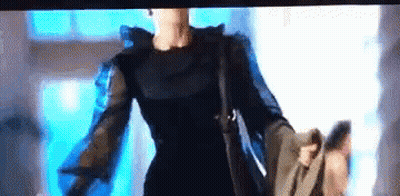 A Woman In A Black Dress Is Holding A Purse And Walking Down A Street . GIF