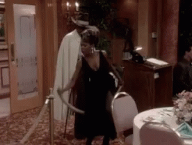 A Woman In A Black Dress Is Holding A Sword In A Room With A Table And Chairs . GIF